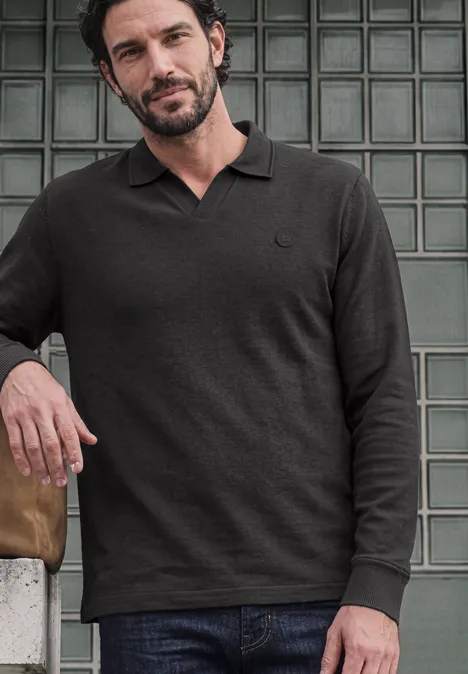 Un homme appuyé sur un mur de briques de verre porte un polo noir à manches longues et un jean.