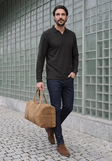 Un homme marche détendu devant une façade vitrée en tenant un sac de voyage.