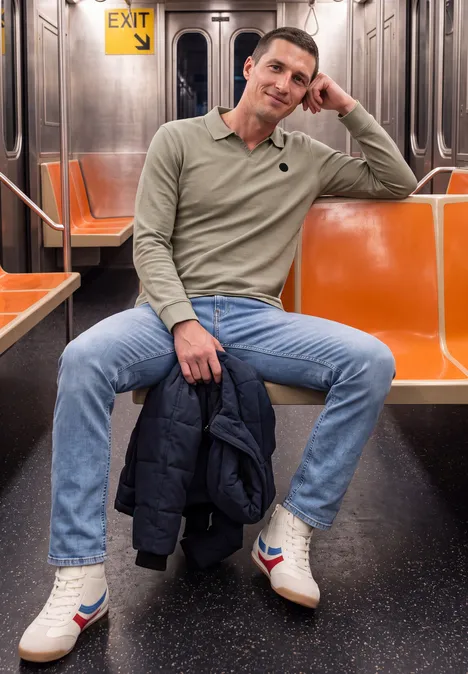 Un homme est assis dans une rame de métro vide, vêtu d’un pull beige, d’un jean et tenant une veste.