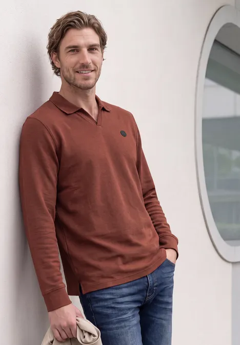 Un homme appuyé sur un mur, portant un pull marron et un jean.