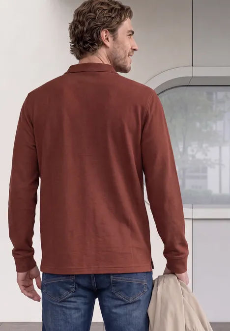 Un homme vu de dos porte un polo marron à manches longues et tient une veste à la main.