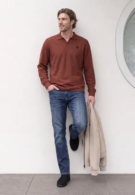 Homme en polo rouge, jean et chaussures noires appuyé contre un mur blanc, tenant une veste beige.