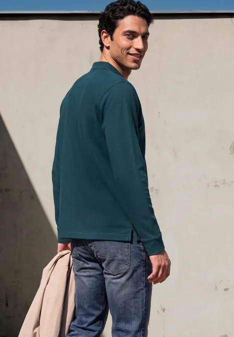 Un homme en jean et polo vert tient une veste beige et regarde par-dessus l'épaule devant un mur clair.