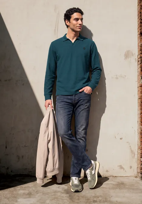 Un homme appuyé contre un mur, porte un polo vert, un jean, des baskets et tient une veste beige.
