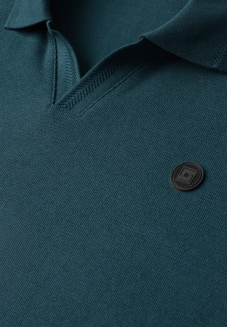 Polo vert à col classique, patte en V texturée et petit badge rond noir sur la poitrine.