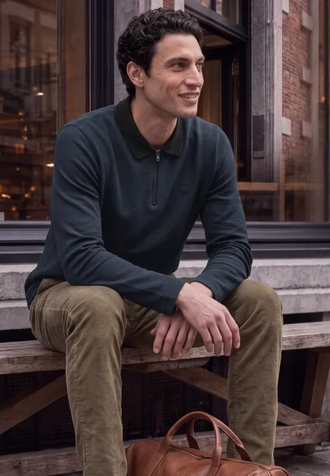 Un homme souriant est assis sur un banc en bois devant un bâtiment, avec un sac en cuir à côté.