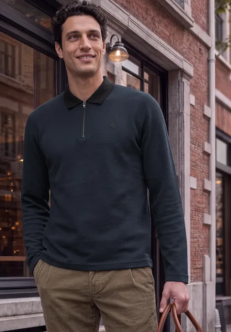 Un homme en polo noir à manches longues et pantalon kaki, tenant un sac devant une façade historique.