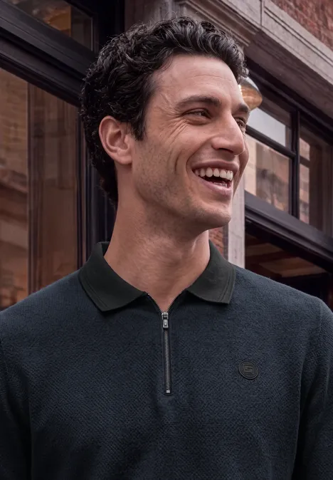 Un homme souriant portant un polo foncé à col et fermeture éclair devant une façade vitrée.