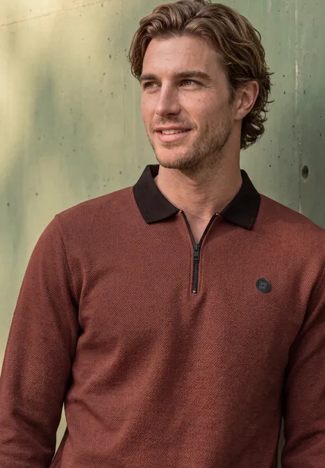 Un homme souriant porte un pull marron à fermeture éclair et col noir, devant un fond métallique vert.