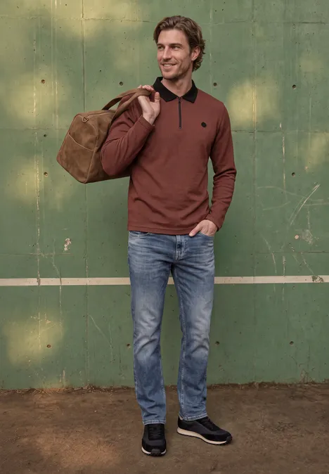 Un homme en jean et pull marron porte un sac de voyage sur l’épaule et se tient devant un mur vert.