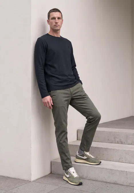 Mann lehnt an weißer Wand, trägt schwarzes Shirt, olivgrüne Hose und grüne Sneaker.