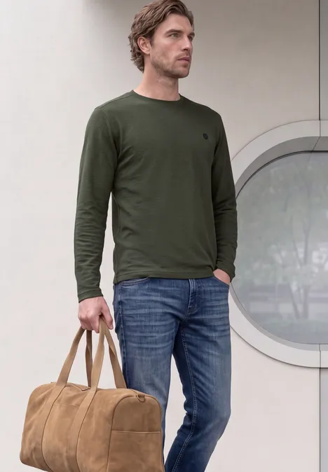 Un homme en jean et t-shirt vert olive à manches longues tient un sac de voyage marron.