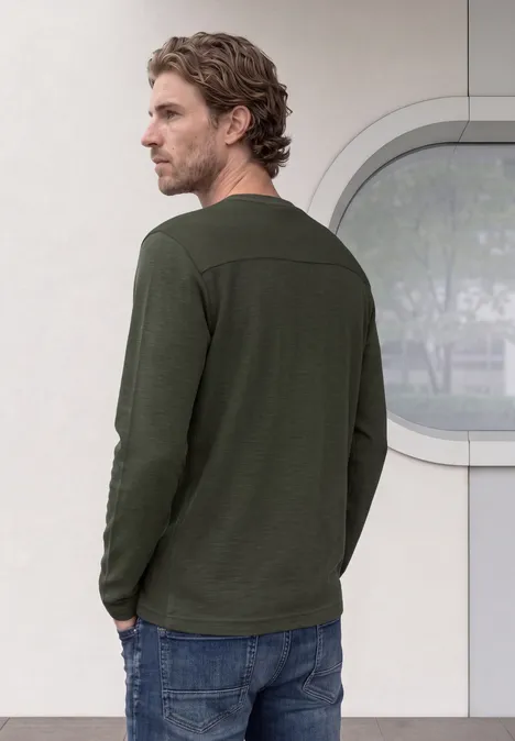 Un homme en t-shirt vert à manches longues et jean, de profil, devant un mur clair.