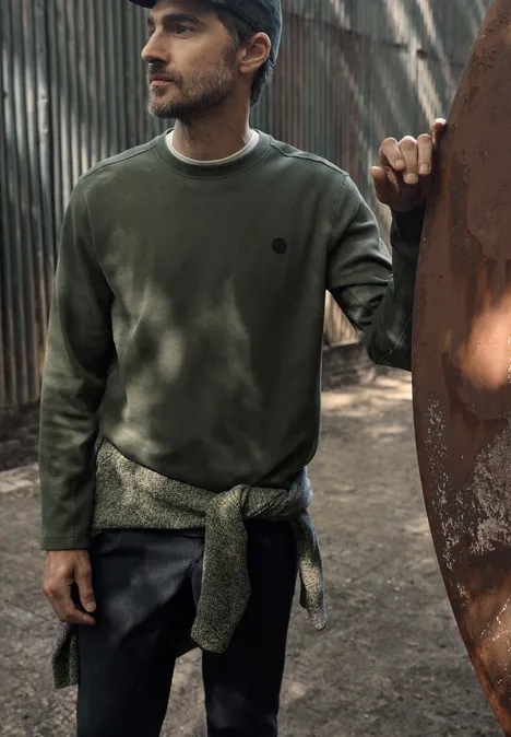 Un homme en sweat vert et casquette, un pull noué autour de la taille, se tient devant une structure métallique en plein air.