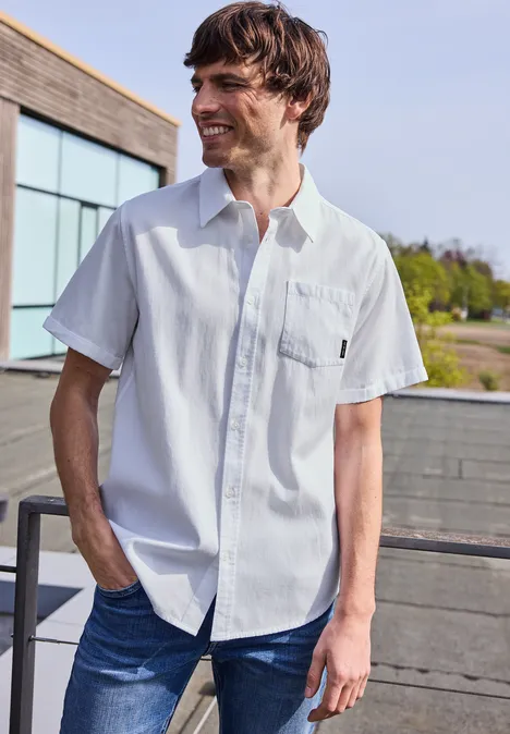 Ein junger Mann trägt weiße Kurzarmhemd und Jeans, steht auf einer Terrasse.