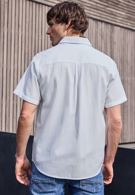 Ein Mann mit kurzärmeligem Hemd und Jeans steht mit dem Rücken zur Kamera vor einer Holzwand.