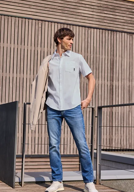 Ein Mann in hellblauem Hemd und Jeans lehnt lässig eine Jacke über die Schulter vor moderner Holz- und Metallfassade.