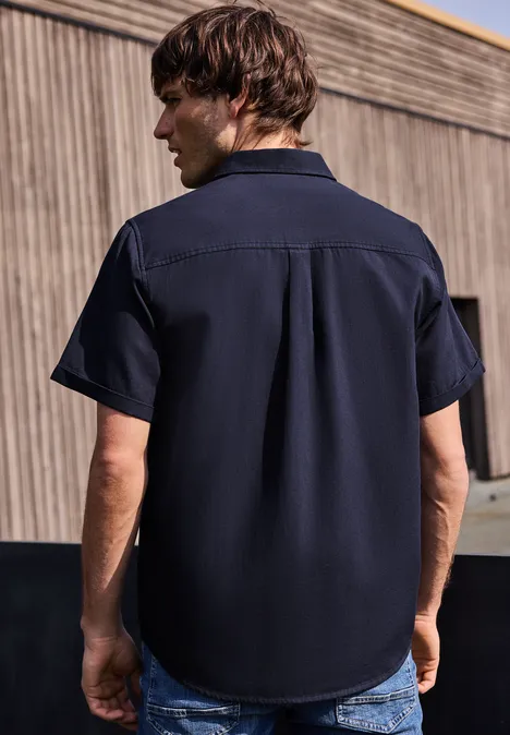 Ein Mann von hinten in einem dunkelblauen Kurzarmhemd steht vor einer Holzfassade im Freien.