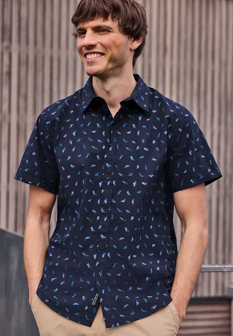 Mann im kurzärmeligen, dunkelblauen Hemd mit hellblauem Muster vor hölzerner Wand.