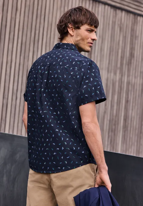 Ein Mann steht vor einer Holzwand, trägt ein navyblaues Hemd mit Muster und eine khakifarbene Hose.