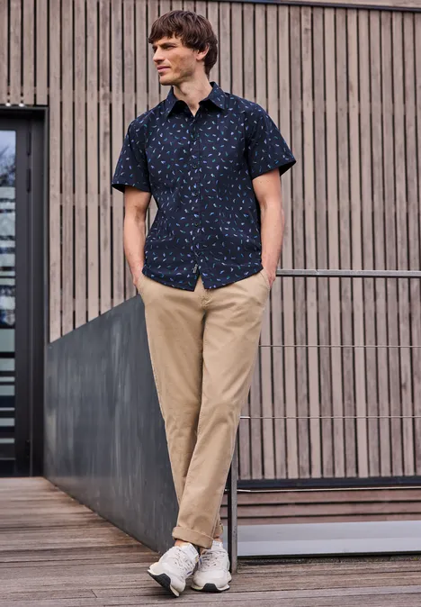 Ein Mann in gemustertem Kurzarmhemd, beiger Hose und Sneakern lehnt locker an einem Geländer vor Holzfassade.