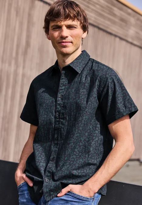 Ein Mann steht lässig vor Holzfassade in einem dunkelblau gemusterten Kurzarmhemd.