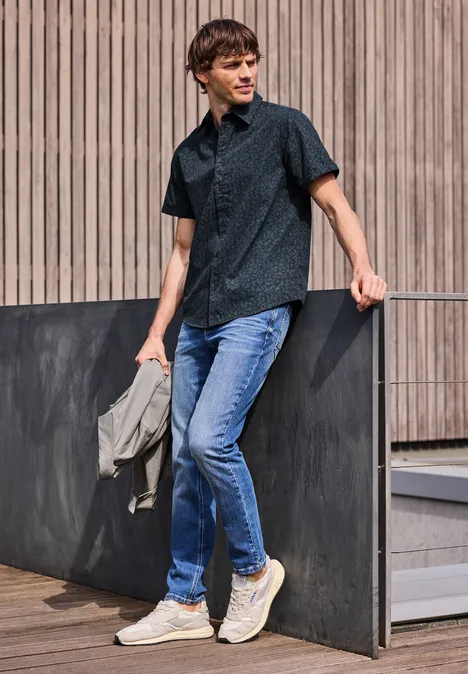 Ein junger Mann lehnt an einer Holzfassade, trägt dunkles Hemd, Jeans und weiße Sneaker.