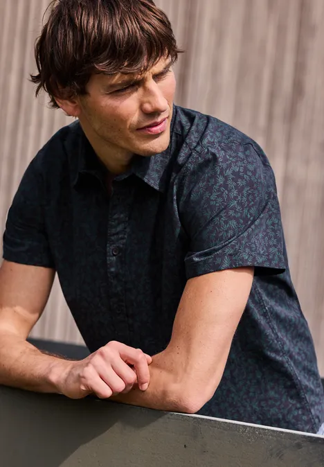 Ein Mann lehnt mit verschränkten Armen auf einem Geländer, trägt ein kurzärmeliges, dunkel gemustertes Hemd vor Holz.