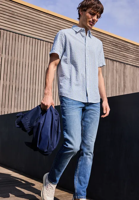 Eine Person trägt ein hellblaues Kurzarmhemd, Jeans und weiße Sneaker, hält eine Jacke und steht im Freien.