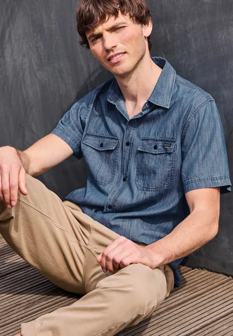 Junger Mann sitzt in legerem Jeanshemd und beigen Chinos vor einer dunklen Wand.