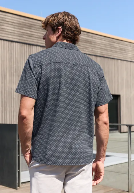 Man van achteren in een kort mouw shirt met klein patroon, modern gebouw en glazen balustrade op de achtergrond.