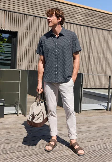 Een man staat op een houten terras, draagt een donker overhemd, lichte broek, sandalen en houdt een tas vast.