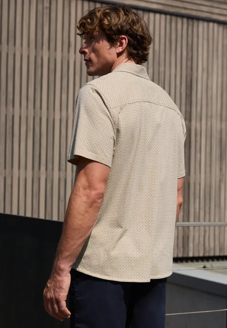 Een man staat buiten met zijn rug naar de camera voor een houten gevel, gekleed in een licht overhemd en donkere broek.