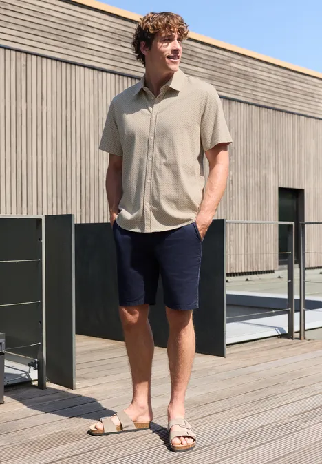 Man in lichtbeige overhemd, donkerblauwe shorts en sandalen, staat ontspannen op een houten terras voor een modern gebouw.