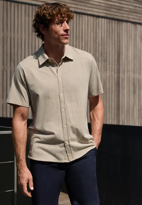 Een man draagt een beige kort mouw shirt en donkere broek voor een houten gevel.