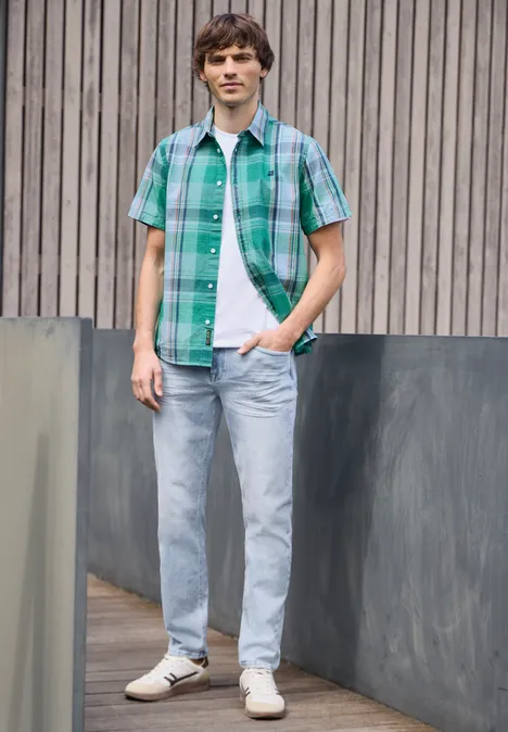 Ein junger Mann in grün kariertem Hemd und Jeans steht lässig vor einer Holzverkleidung.