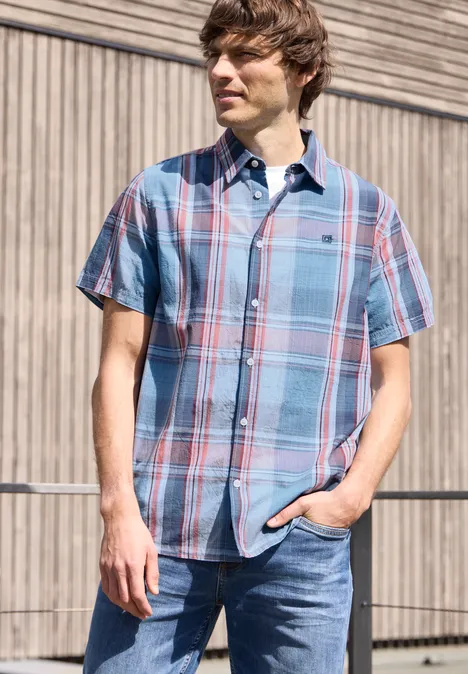 Ein Mann steht vor einer Holzwand, trägt kariertes Kurzarmhemd und Jeans, eine Hand in der Hosentasche.