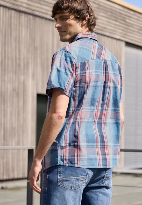 Ein Mann mit kariertem Hemd und Jeans steht vor einem Holzgebäude und schaut weg.