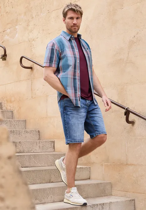 Ein Mann in Sommeroutfit steht auf einer Steintreppe und lehnt an einem Handlauf vor einer hellen Wand.