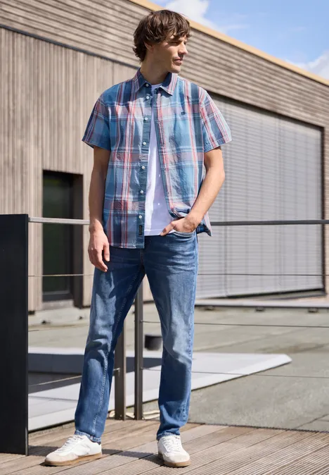 Ein junger Mann lehnt entspannt am Geländer vor einem modernen Gebäude, trägt karierte Hemd und Jeans.