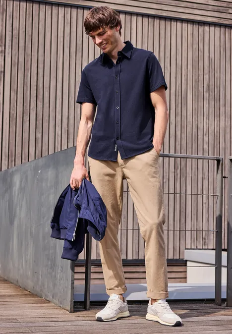 Junger Mann in navyblauem Hemd, hellen Chinos und weißen Sneakern, hält eine blaue Jacke und steht im Freien.