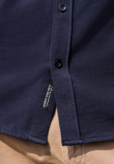 Dunkelblaues Hemd mit Knöpfen, seitlichem Schlitz und Labeldetail, auf beiger Hose.