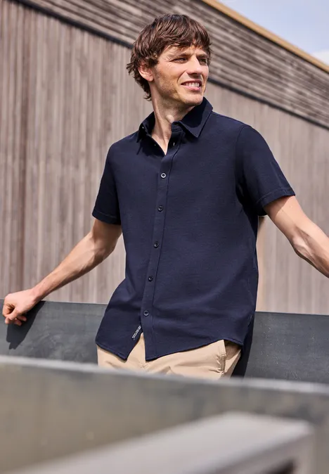 Mann in dunkelblauem Kurzarmhemd lehnt an Metallbrüstung vor einer Holzfassade.