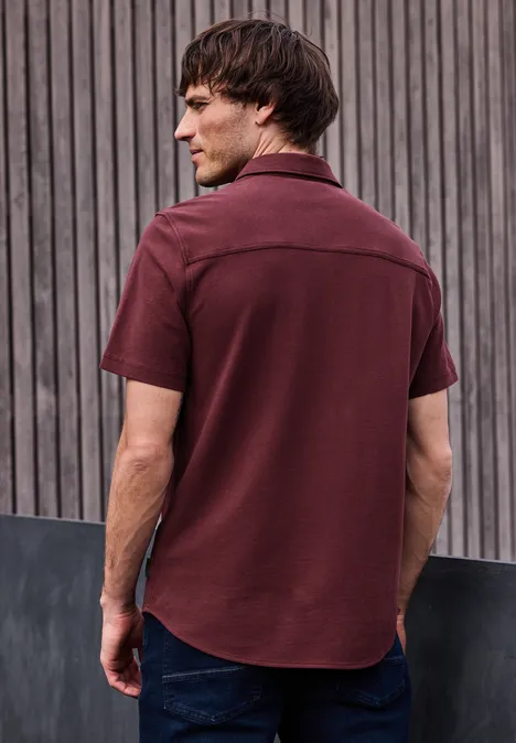 Ein Mann steht mit dem Rücken zur Kamera, trägt ein weinrotes Hemd und Jeans vor einer Holzlattenwand.