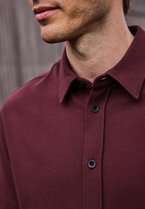 Ein Mann trägt ein bordeauxrotes Poloshirt mit Kragen und Knopfleiste, dunkle Knöpfe, Holzwand im Hintergrund.