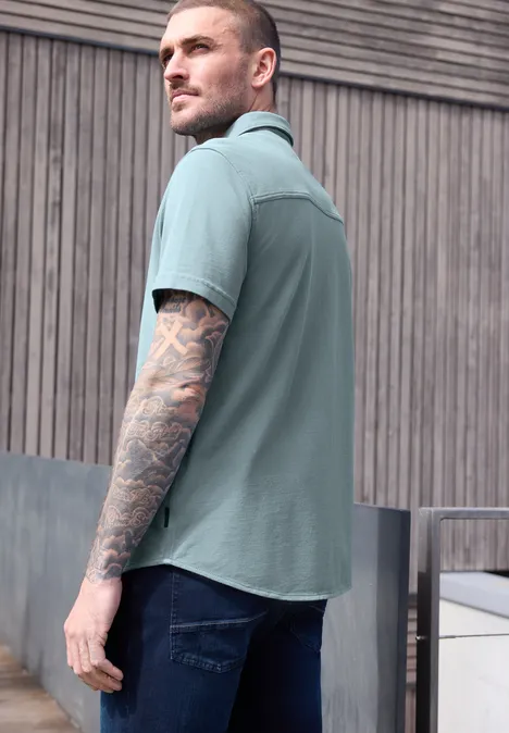 Ein Mann im mintgrünen Kurzarmhemd mit Arm-Tätowierungen steht seitlich vor einer Holzwand.