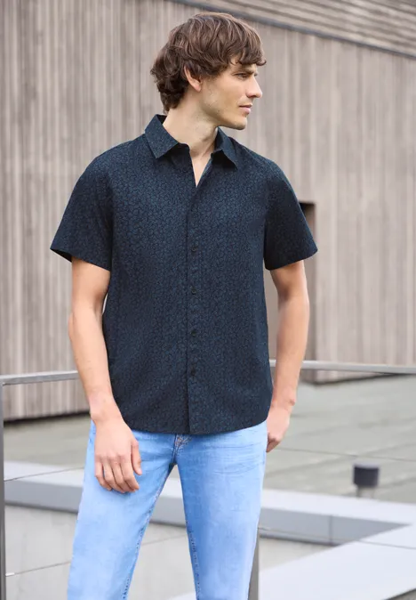 Ein Mann in dunkelblauem Kurzarmhemd und hellblauen Jeans steht vor einer Holzfassade.