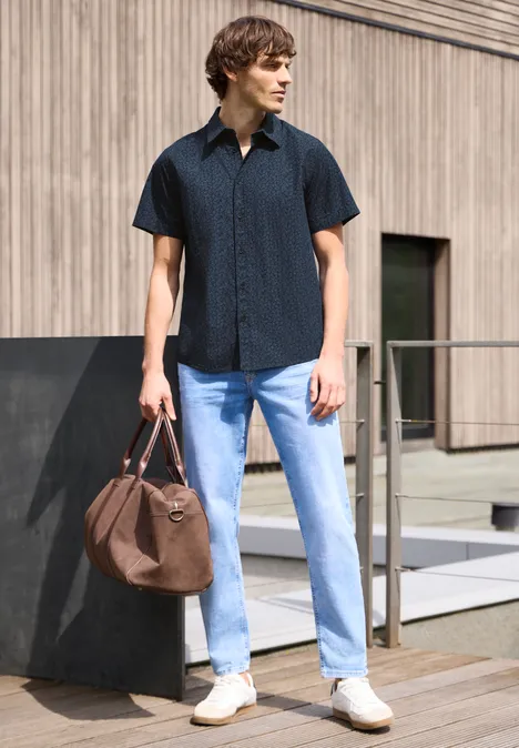 Ein Mann steht vor Gebäude, trägt schwarzes Hemd, helle Jeans, weiße Sneaker und hält eine braune Tasche.