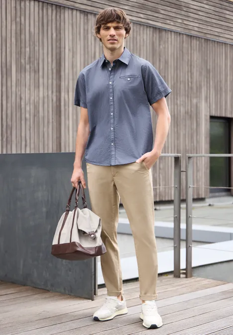 Mann in dunkelblauem Hemd, beiger Hose und Sneakers hält eine Tasche vor einer Holzfassade.