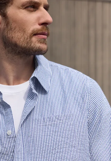 Profilaufnahme eines bärtigen Mannes mit weißem T-Shirt und blau-weiß gestreiftem Hemd vor hellem Hintergrund